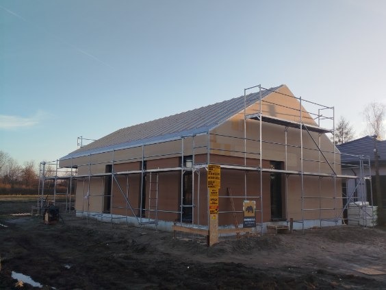 Budowa domu jednorodzinnego w trakcie ocieplania, z widocznymi rusztowaniami i częściowo ocieploną elewacją. Ujęcie z boku, w tle błękitne niebo i sąsiednie budynki.