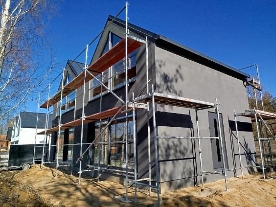 Nowoczesny dom z szarą elewacją w trakcie budowy, otoczony rusztowaniami. Widoczne duże okna i fragmenty innych budynków w tle, błękitne niebo.