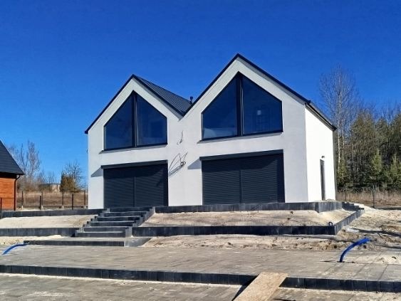 Biały, nowoczesny dom z dwuspadowym dachem i dużymi oknami, z szarymi roletami garażowymi. Uwagę przyciągają schody prowadzące do wejścia i częściowo wykończony teren wokół.