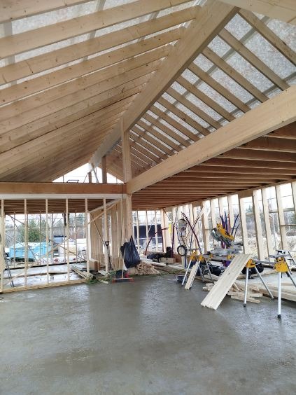Wnętrze w trakcie budowy: drewniana konstrukcja dachu z widocznymi belkami, ściany szkieletowe, narzędzia budowlane i świeża wylewka podłogowa. Surowy stan, etap prac.