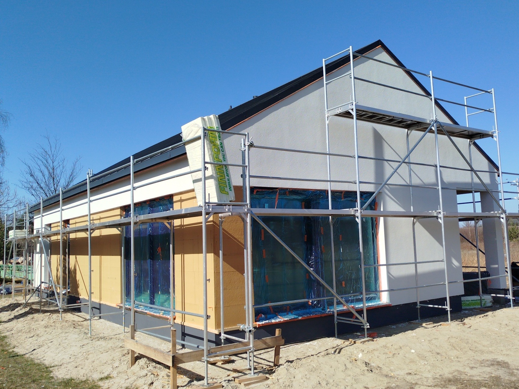 Budowa domu szkieletowego w Nisko na etapie ocieplania, widoczne rusztowania i materiały izolacyjne Finna Foam. Jasne tło nieba.