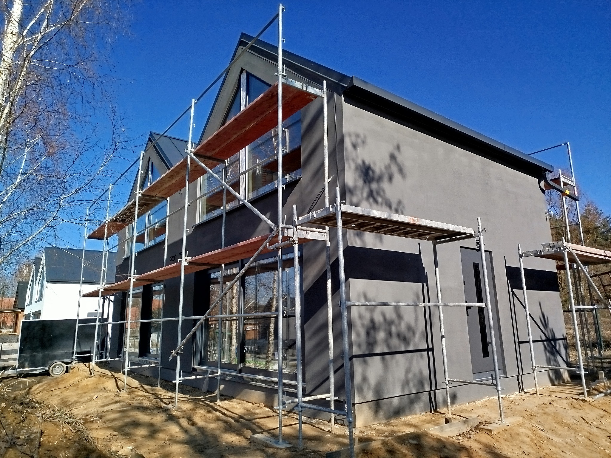 Nowoczesny dom z szarą elewacją w trakcie budowy, otoczony rusztowaniami. Widoczne duże okna i fragment przyczepy. Błękitne niebo w tle.