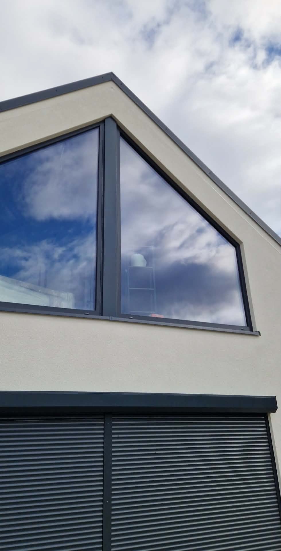 Fragment elewacji domu z trójkątnym oknem, w którym odbija się niebo. Roleta zewnętrzna w kolorze antracytowym. Jasny tynk.