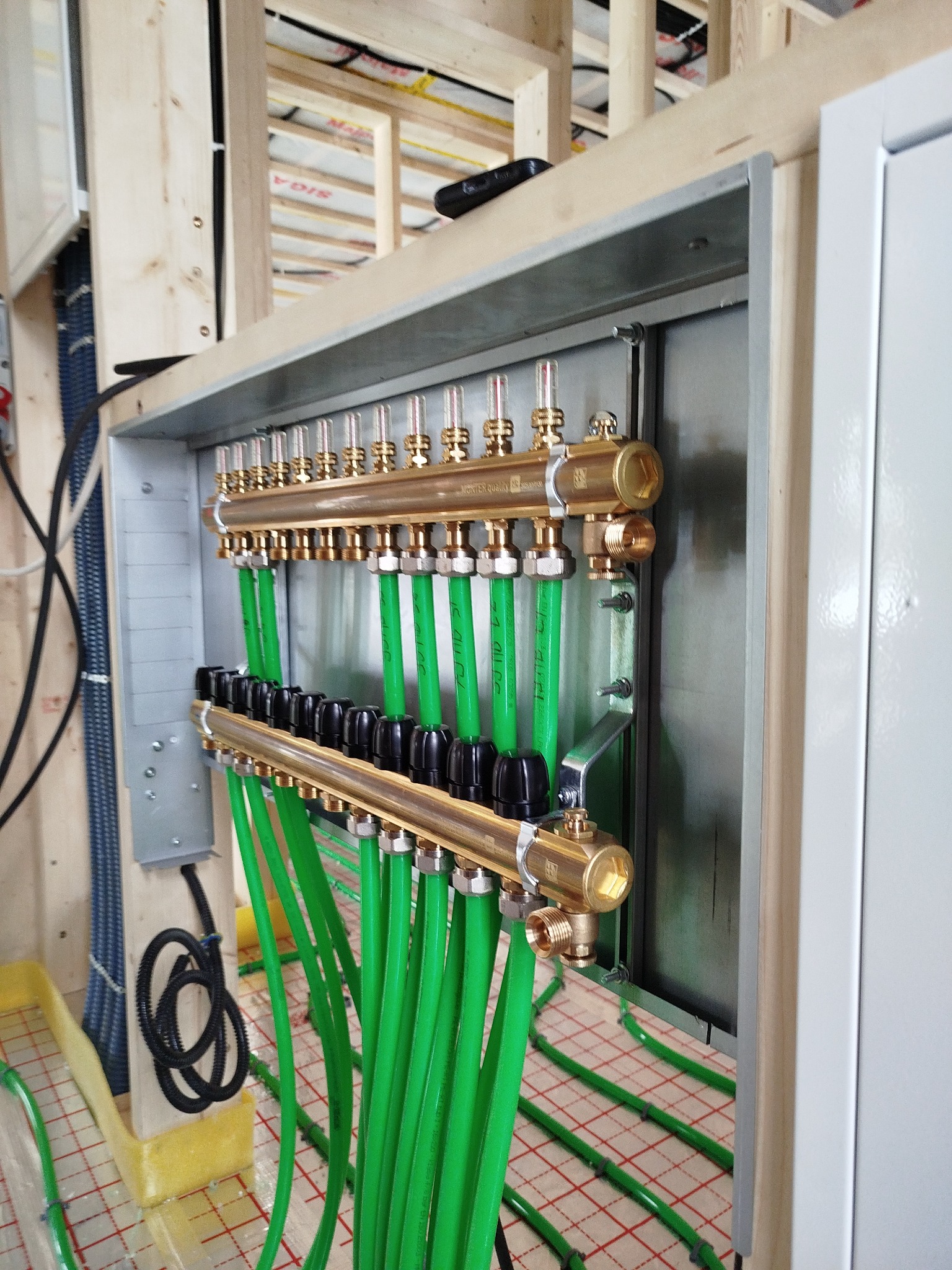 Rozdzielacz ogrzewania podłogowego z zielonymi rurami w drewnianej konstrukcji domu, widoczne elementy instalacji i podłoże z siatką. Detal montażu w surowym stanie.