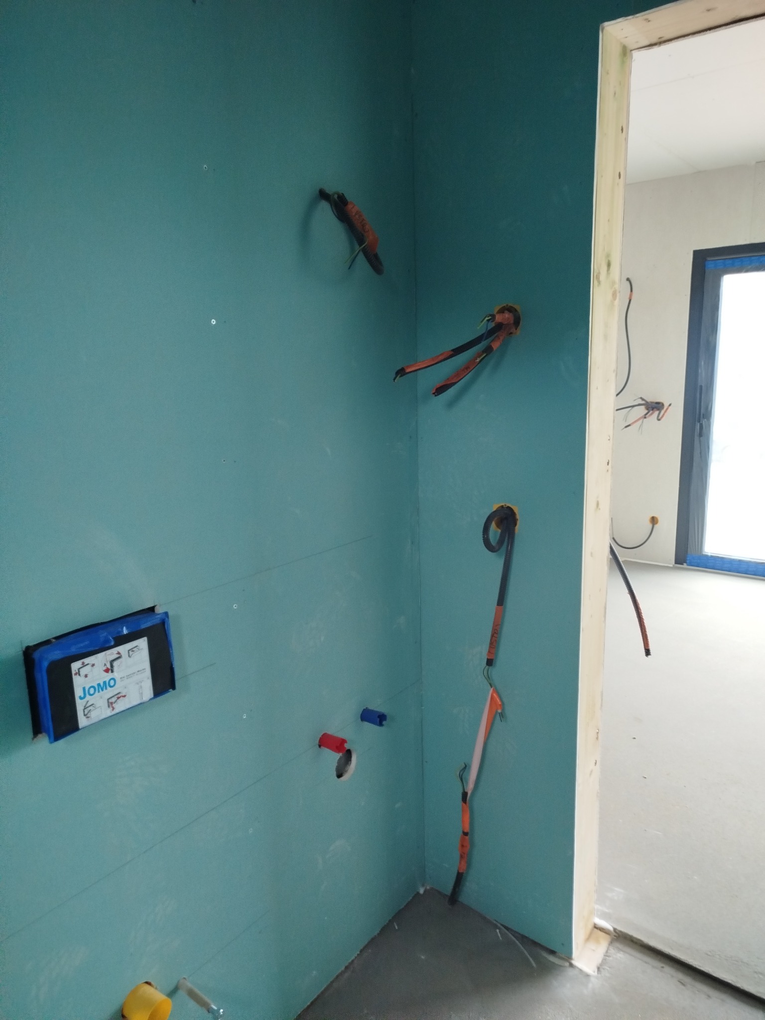 Niebieska ściana z instalacją elektryczną i wodną wewnątrz domu w trakcie budowy. Widoczne kable, rury i puszka JOMO. Surowy stan wykończenia.