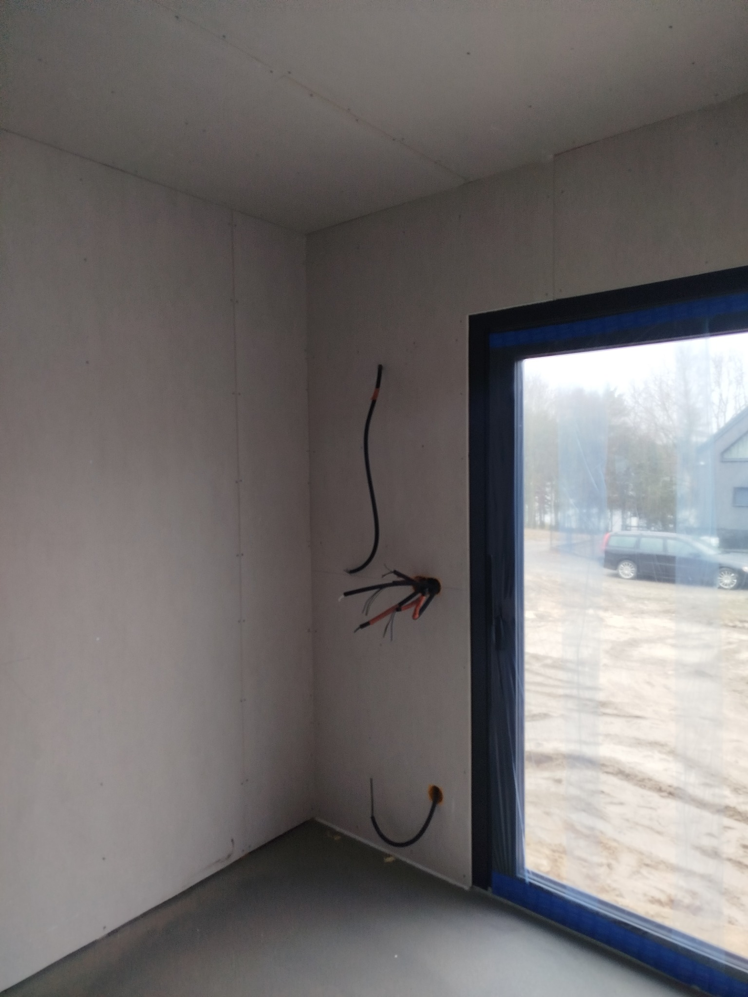 Wnętrze w trakcie budowy domu szkieletowego: ściany z płyt, okno z niebieską folią ochronną, wystające przewody elektryczne. Widok na zewnątrz.