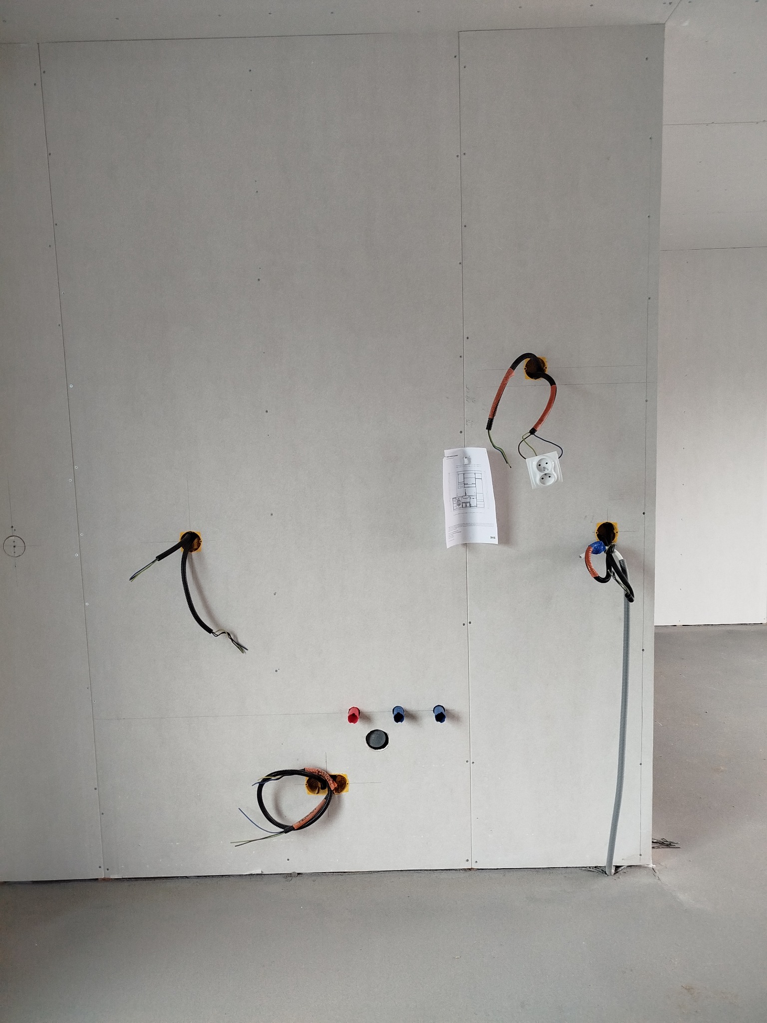 Ściana z płyt gipsowo-kartonowych w trakcie montażu instalacji elektrycznej i hydraulicznej. Widoczne kable, gniazdka, rury i schemat instalacji.