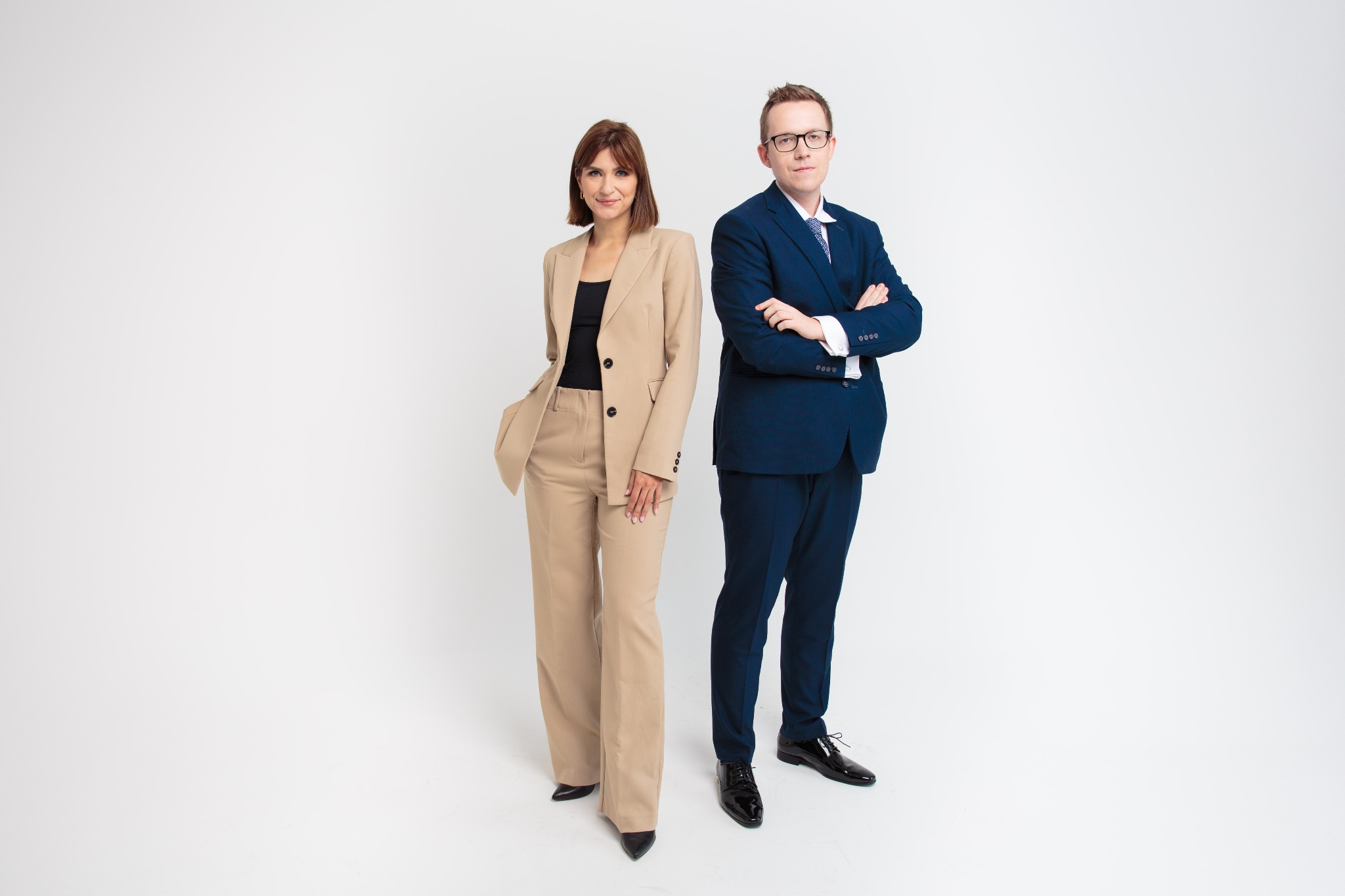Profesjonalny portret zespołowy: kobieta w beżowym garniturze i mężczyzna w granatowym garniturze, oboje stoją na białym tle, uśmiechają się i patrzą w kamerę.