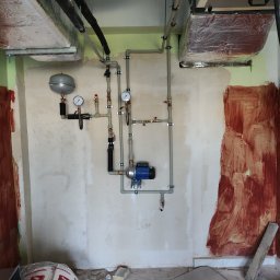 AIR-KAN Marcin Szura Instalacje sanitarne - Instalacja grzewcza z pompą obiegową, manometrami i zbiornikiem wyrównawczym, zamontowana na surowej ścianie w trakcie remontu, widoczne rury izolowane na suficie i materiały budowlane na podłodze.