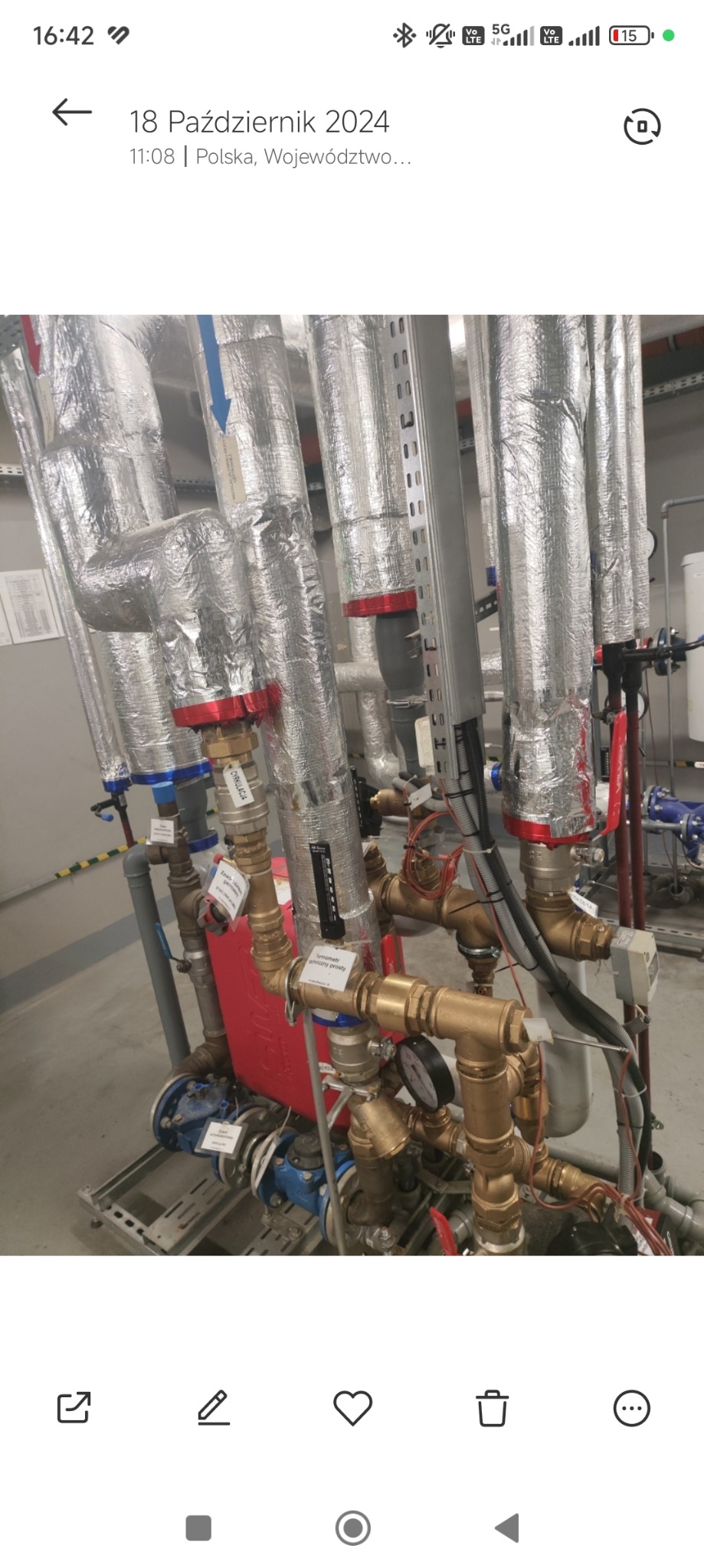 Złożona instalacja hydrauliczna z rurami izolowanymi folią aluminiową, widoczne zawory, manometry i etykiety opisowe, całość w pomieszczeniu technicznym.