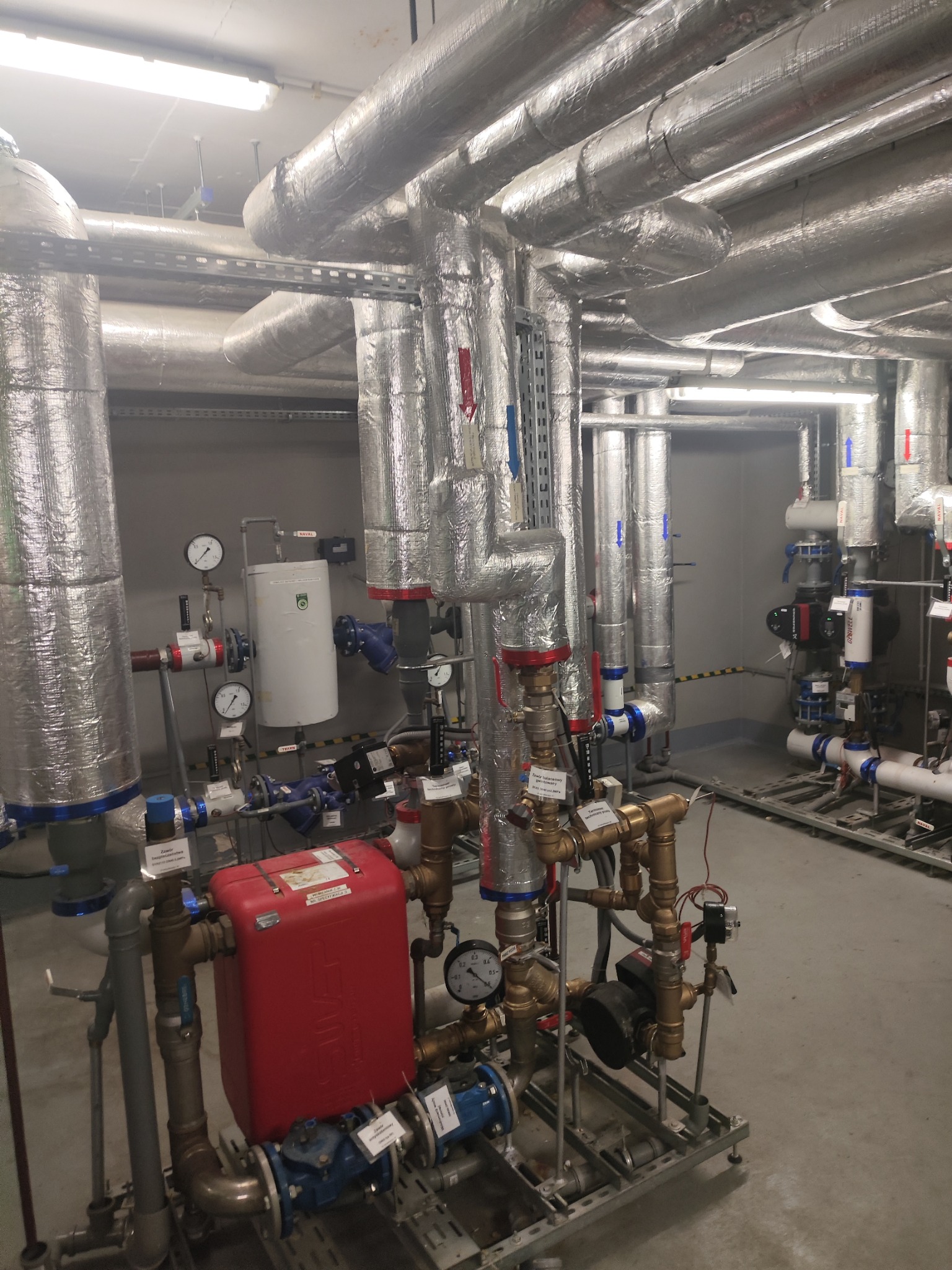 Złożona instalacja rur i zaworów w pomieszczeniu technicznym, rury zaizolowane folią aluminiową, manometry i pompy widoczne, oświetlenie jarzeniowe sufitowe.