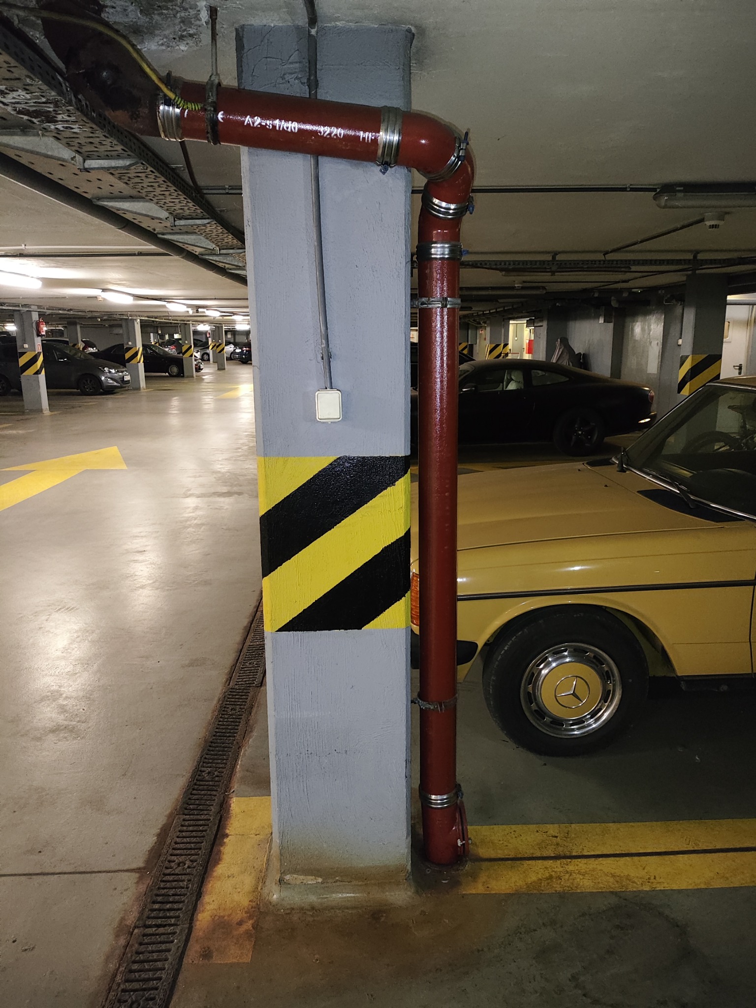 Czerwona rura instalacyjna z obejmami biegnąca wzdłuż szarego słupa w garażu podziemnym, żółto-czarne pasy ostrzegawcze na słupie, żółty samochód Mercedes w tle.
