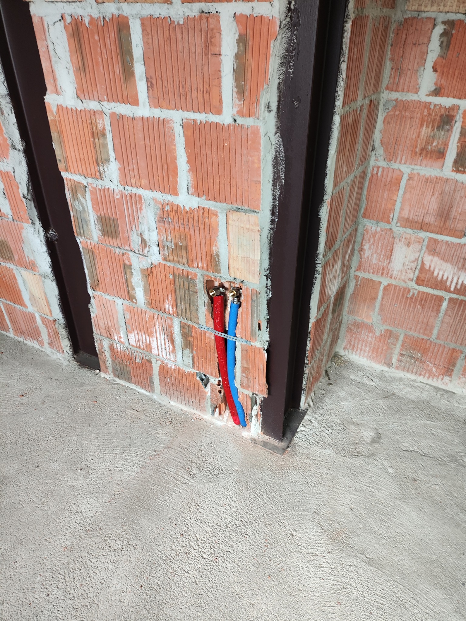 Wystające z muru dwa rurki, czerwona i niebieska, instalacja w trakcie wykonywania w Gorlicach, widok na surową cegłę i betonową podłogę.