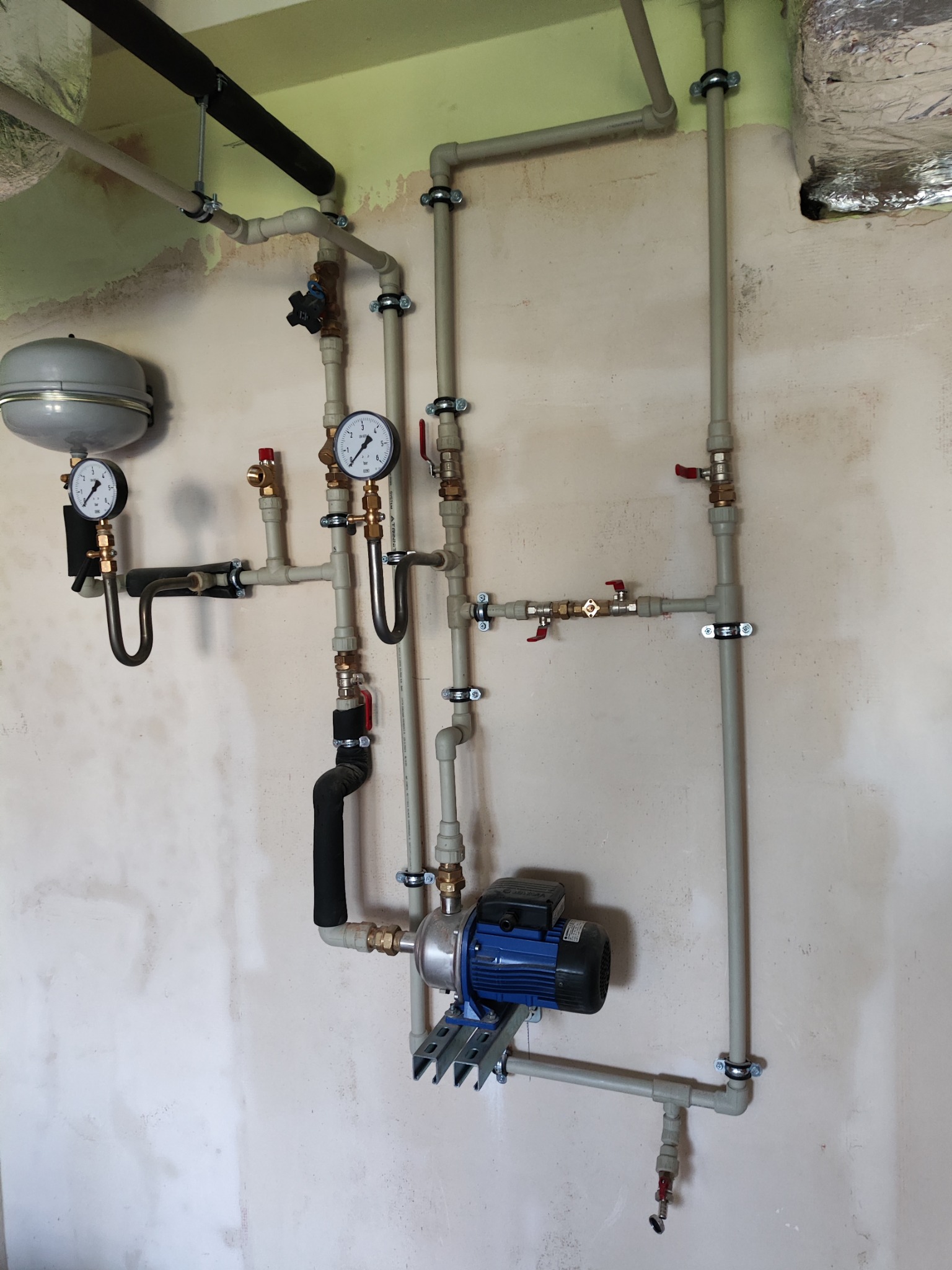 Kompleksowa instalacja hydrauliczna z pompą obiegową, manometrami i rurami z tworzywa sztucznego zamocowanymi na ścianie w surowym stanie.
