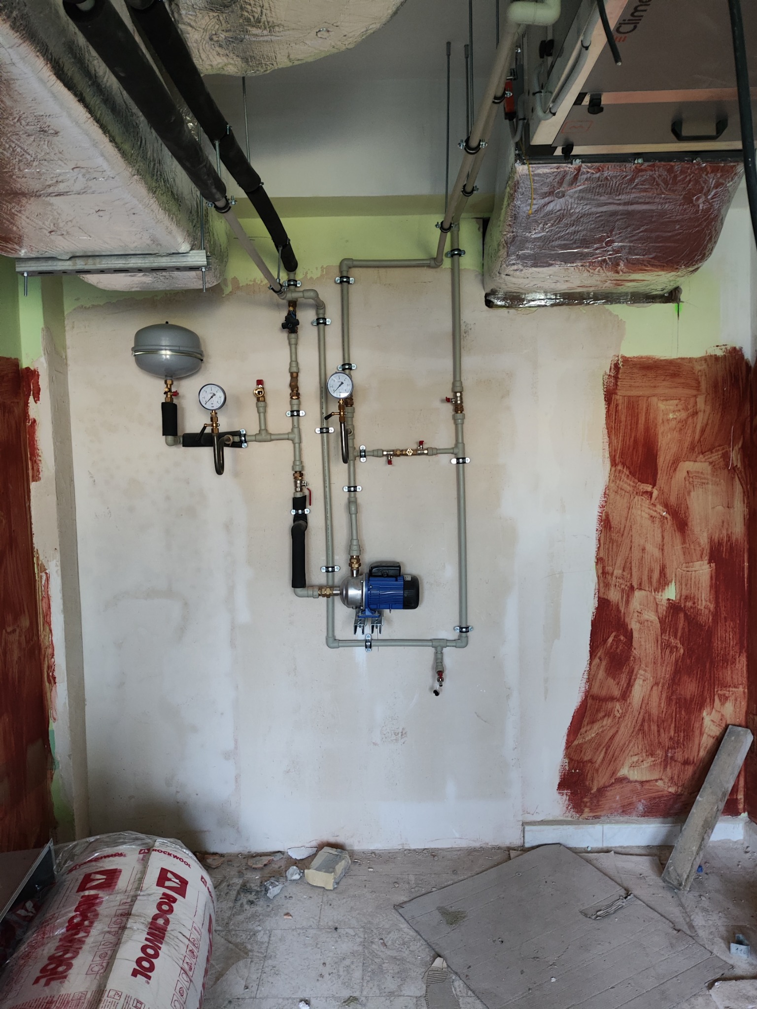 Instalacja grzewcza z pompą obiegową, manometrami i zbiornikiem wyrównawczym, zamontowana na surowej ścianie w trakcie remontu, widoczne rury izolowane na suficie i materiały budowlane na podłodze.