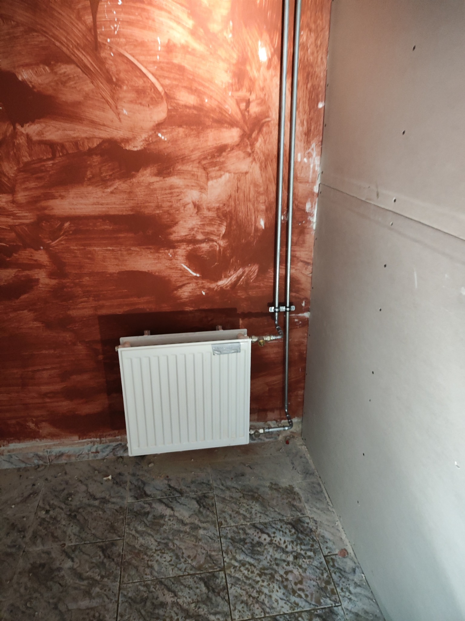 Biały grzejnik panelowy zamontowany na ścianie z niedokończonym malowaniem, widoczne rury doprowadzające wodę i płyty kartonowo-gipsowe na ścianie obok.