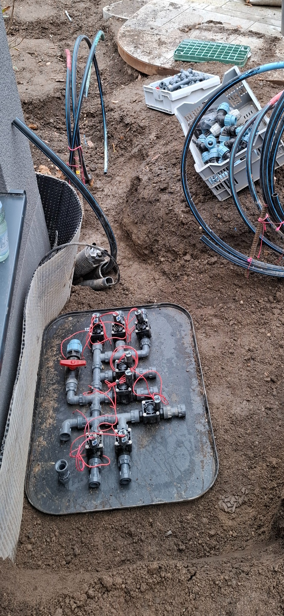 Rozłożony na czarnej tacy system zaworów i rur z czerwonymi przewodami, przygotowany do montażu systemu nawadniania w wykopie z widocznymi fragmentami rur, skrzynek złączek i ziemi.