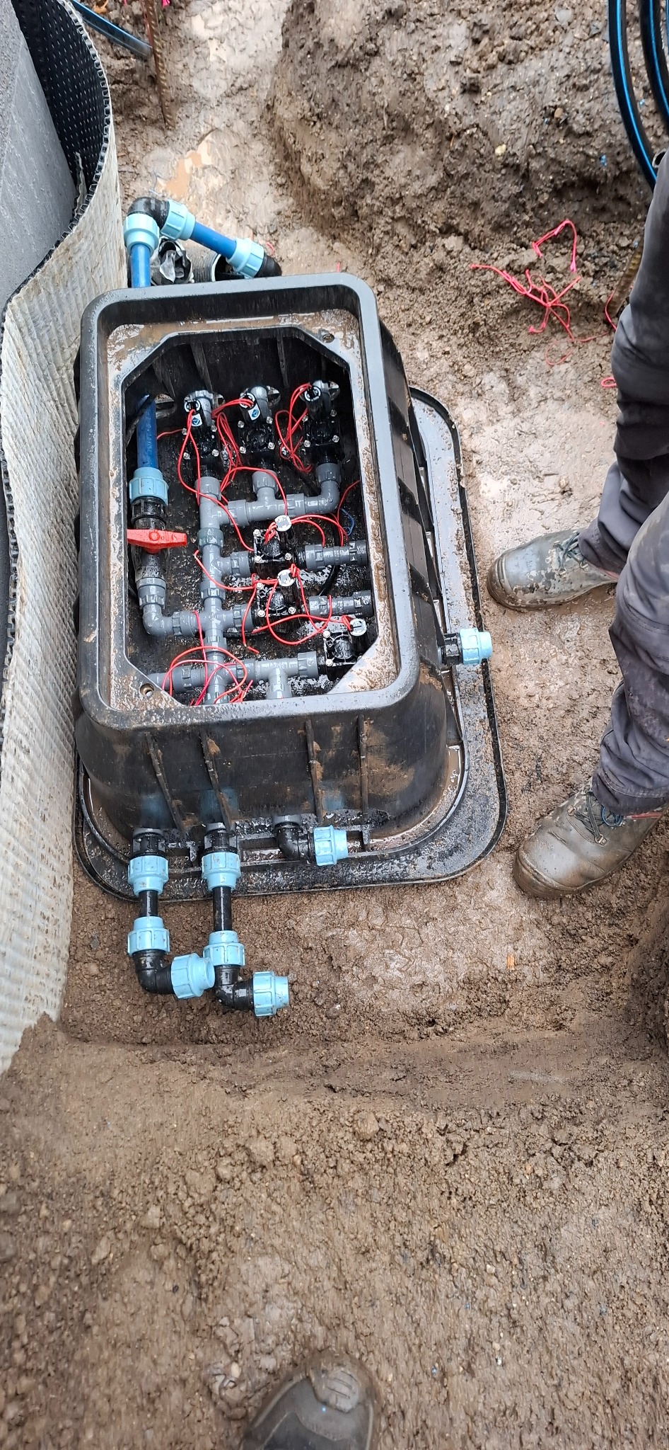 Otwarta skrzynka z zaworami i przewodami instalacji nawadniającej w wykopie, widoczne elementy sterujące i połączenia elektryczne, obok ziemia i fragment buta pracownika.