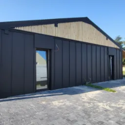 Nowoczesny budynek z czarną blachą na elewacji i drewnianymi elementami wykończeniowymi, widok z zewnątrz na kostkę brukową i fragment ogrodu.