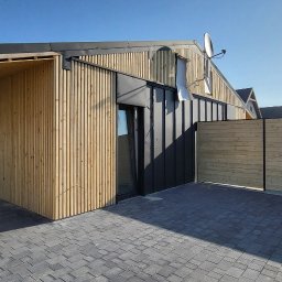 Krzysztof Tomala - Nowoczesny dom z drewnianą elewacją, czarnymi akcentami i brukowanym podjazdem, widok z zewnątrz w słoneczny dzień.