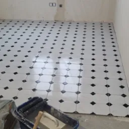 Układanie białych płytek podłogowych z czarnymi akcentami w romby, widoczny kubeł z zaprawą i narzędzia glazurnicze, surowe ściany w tle.