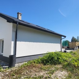 Krzysztof Tomala - Ocieplony i pomalowany na biało dom jednorodzinny w Żorach, widok z boku na elewację i fragment dachu, z widocznym fragmentem ogrodu i kontenerem w tle.