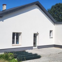Krzysztof Tomala - Nowo otynkowany budynek jednorodzinny z szarą podmurówką i dachem, widoczne rolki zielonej siatki ogrodzeniowej, w tle drzewa i maszyny budowlane.