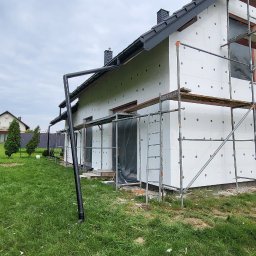 Krzysztof Tomala - Dom jednorodzinny w trakcie ocieplania styropianem, widoczne rusztowania i pracownik w czerwonej bluzie na górze, zielony trawnik i drzewa w tle.