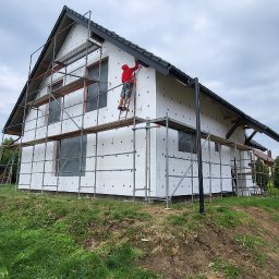 Krzysztof Tomala - Dom jednorodzinny w trakcie ocieplania: biały styropian na elewacji, rusztowania, pracownik w czerwonej bluzie na drabinie, widok z dołu na tle pochmurnego nieba.