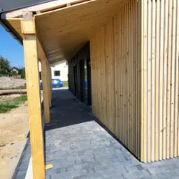 Nowoczesna elewacja budynku z drewnianymi panelami pionowymi i zadaszeniem z surowego drewna, kostka brukowa na podłożu, błękitne niebo w tle.