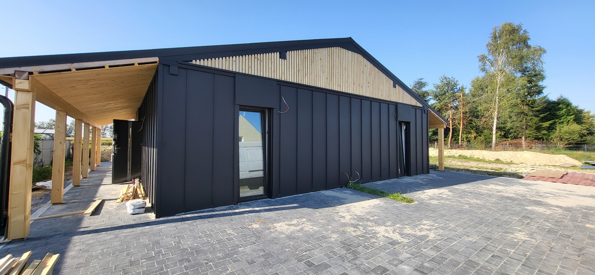 Nowoczesny budynek z czarną blachą na elewacji i drewnianymi elementami wykończeniowymi, widok z zewnątrz na kostkę brukową i fragment ogrodu.