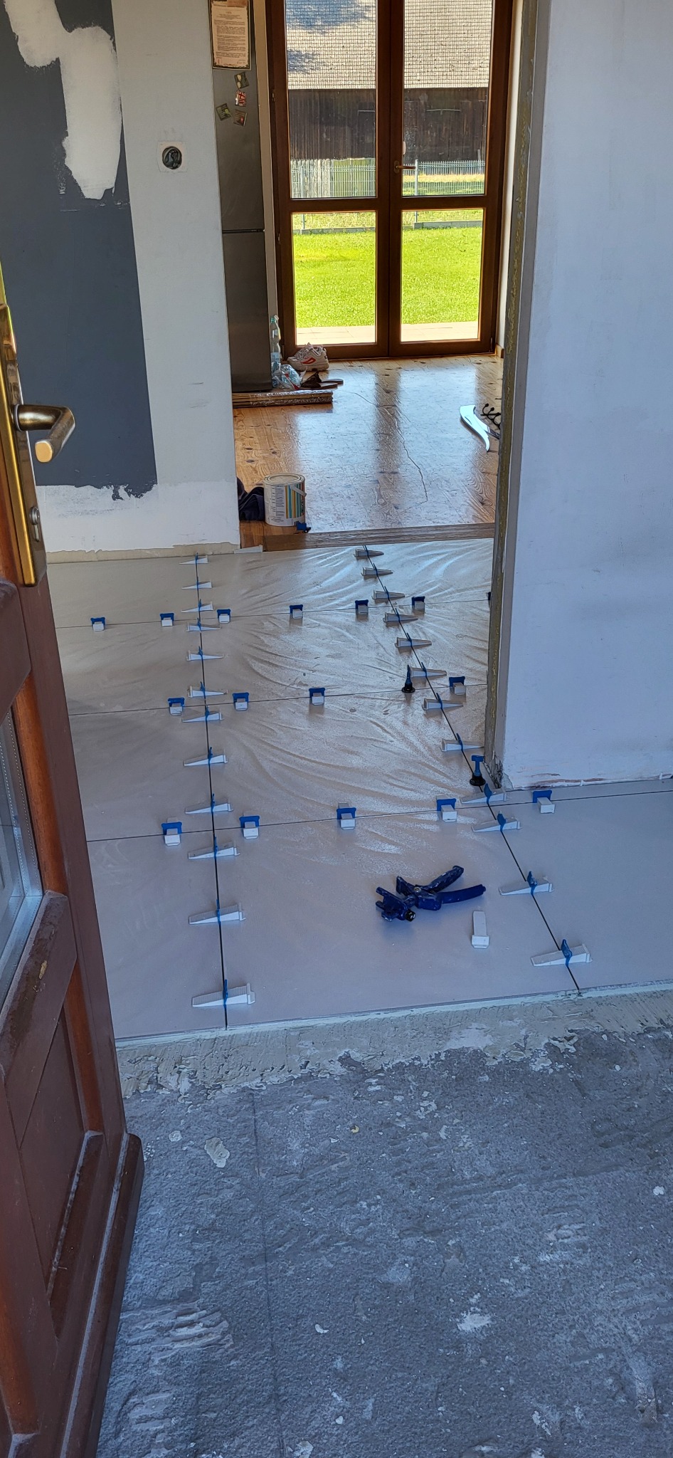 Układanie płytek podłogowych w trakcie remontu, widoczne narzędzia do poziomowania i krzyżyki dystansowe, przez otwarte drzwi widoczna drewniana podłoga w sąsiednim pomieszczeniu.