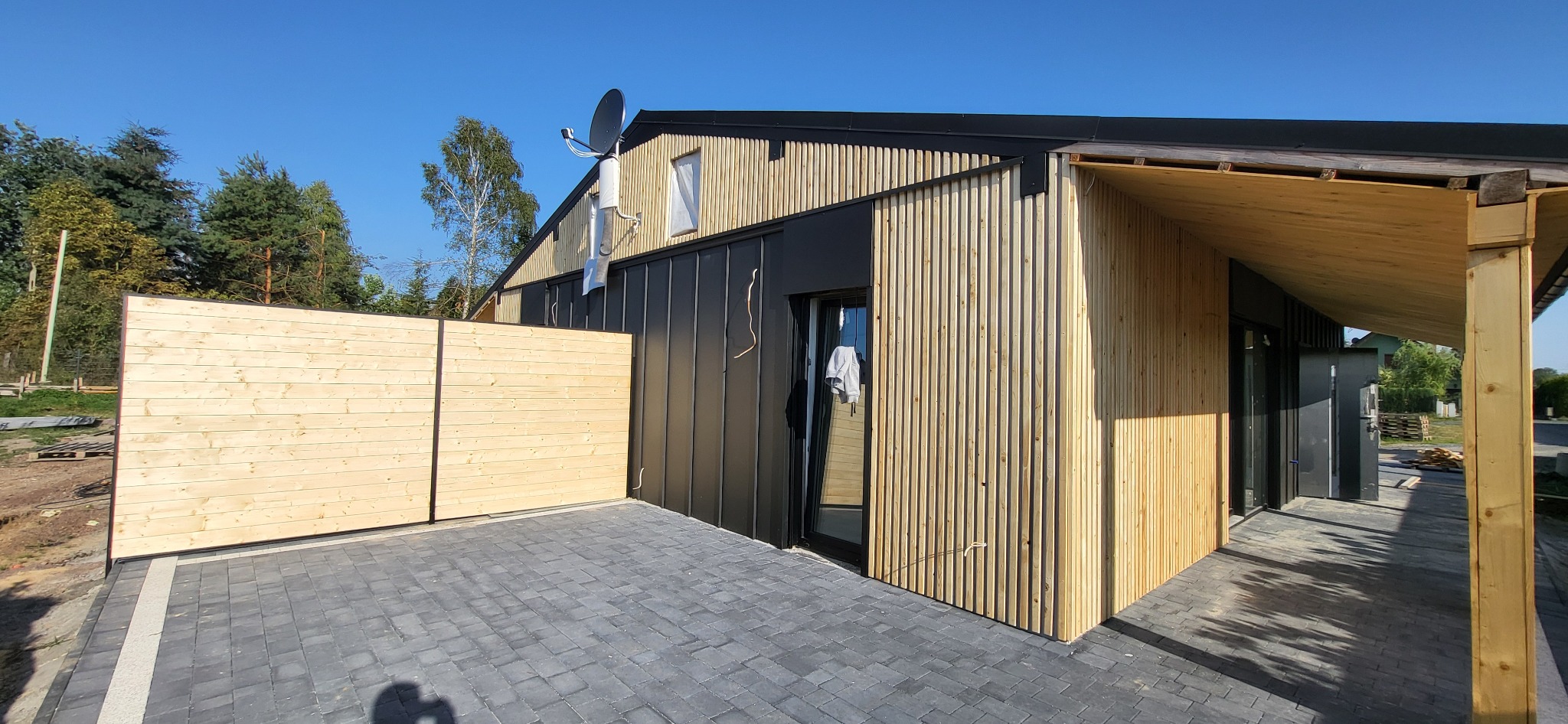 Nowoczesny dom z elewacją z jasnego drewna i czarnymi panelami, widok z zewnątrz, z drewnianym ogrodzeniem i utwardzonym podjazdem z kostki brukowej.