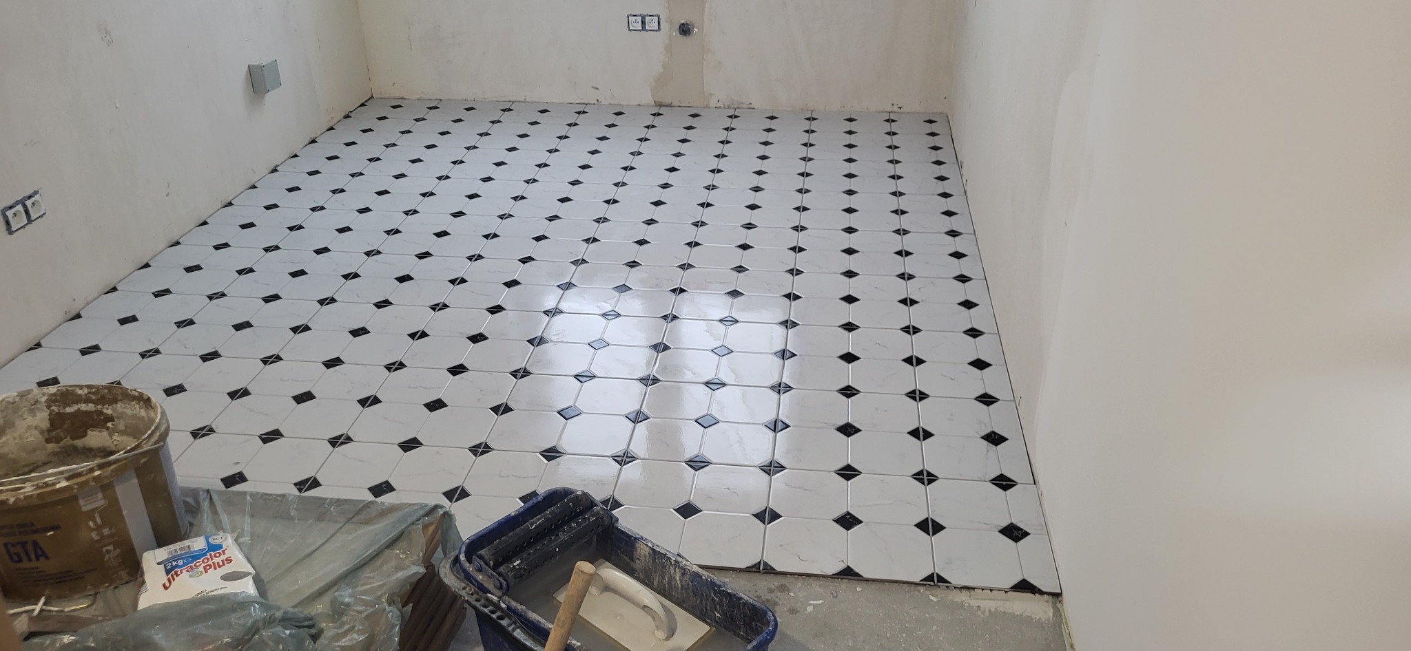Układanie białych płytek podłogowych z czarnymi akcentami w romby, widoczny kubeł z zaprawą i narzędzia glazurnicze, surowe ściany w tle.