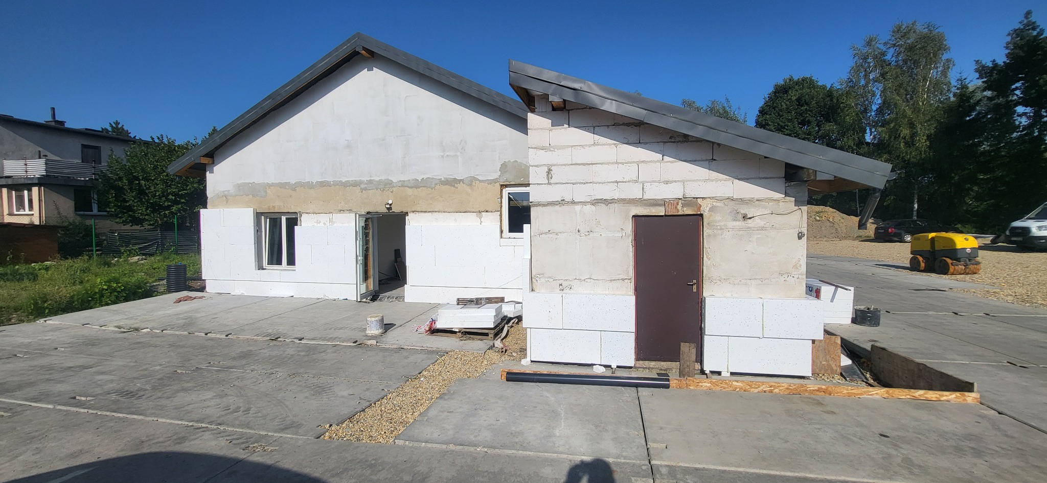 Dom jednorodzinny w trakcie ocieplania elewacji styropianem, widoczne białe płyty izolacyjne, surowy betonowy podjazd, częściowo otynkowana fasada, ciemne okna i drzwi, maszyna budowlana w tle.