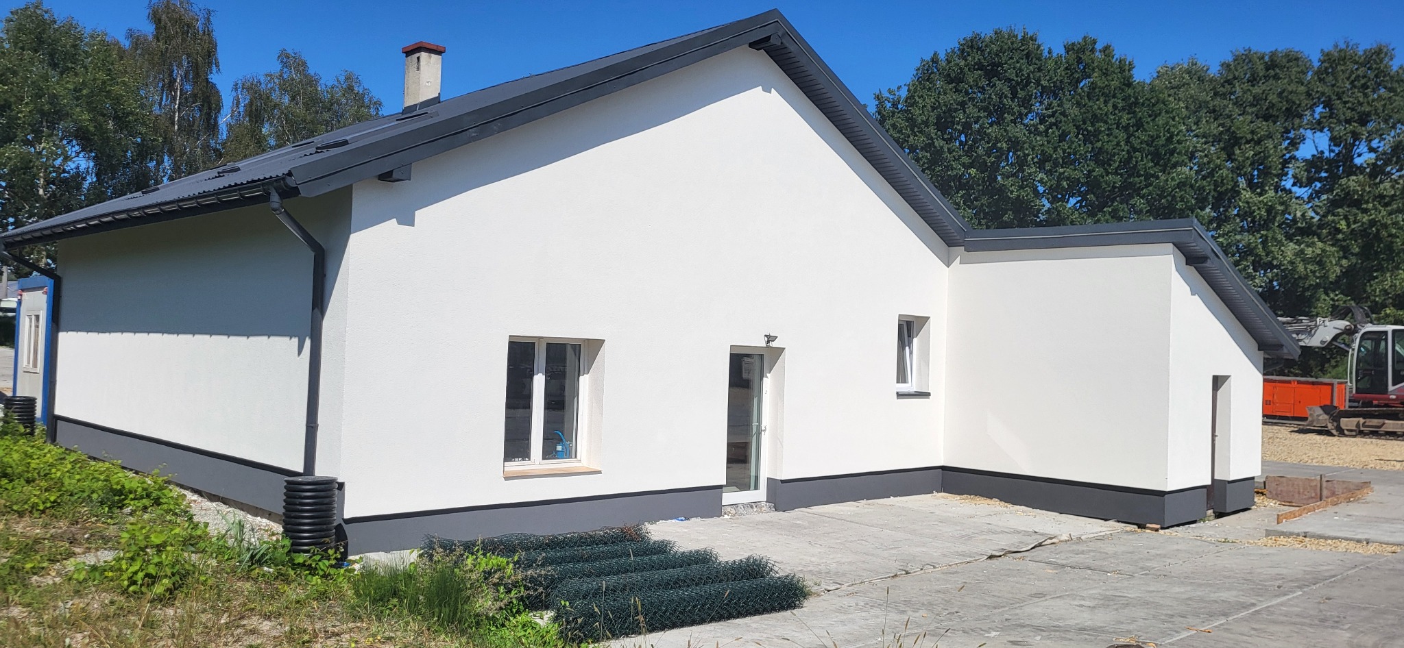 Nowo otynkowany budynek jednorodzinny z szarą podmurówką i dachem, widoczne rolki zielonej siatki ogrodzeniowej, w tle drzewa i maszyny budowlane.