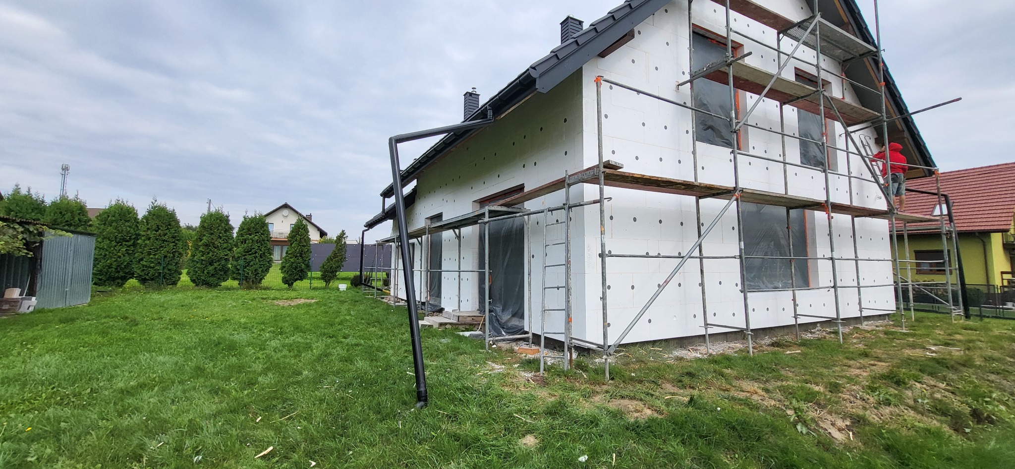 Dom jednorodzinny w trakcie ocieplania styropianem, widoczne rusztowania i pracownik w czerwonej bluzie na górze, zielony trawnik i drzewa w tle.