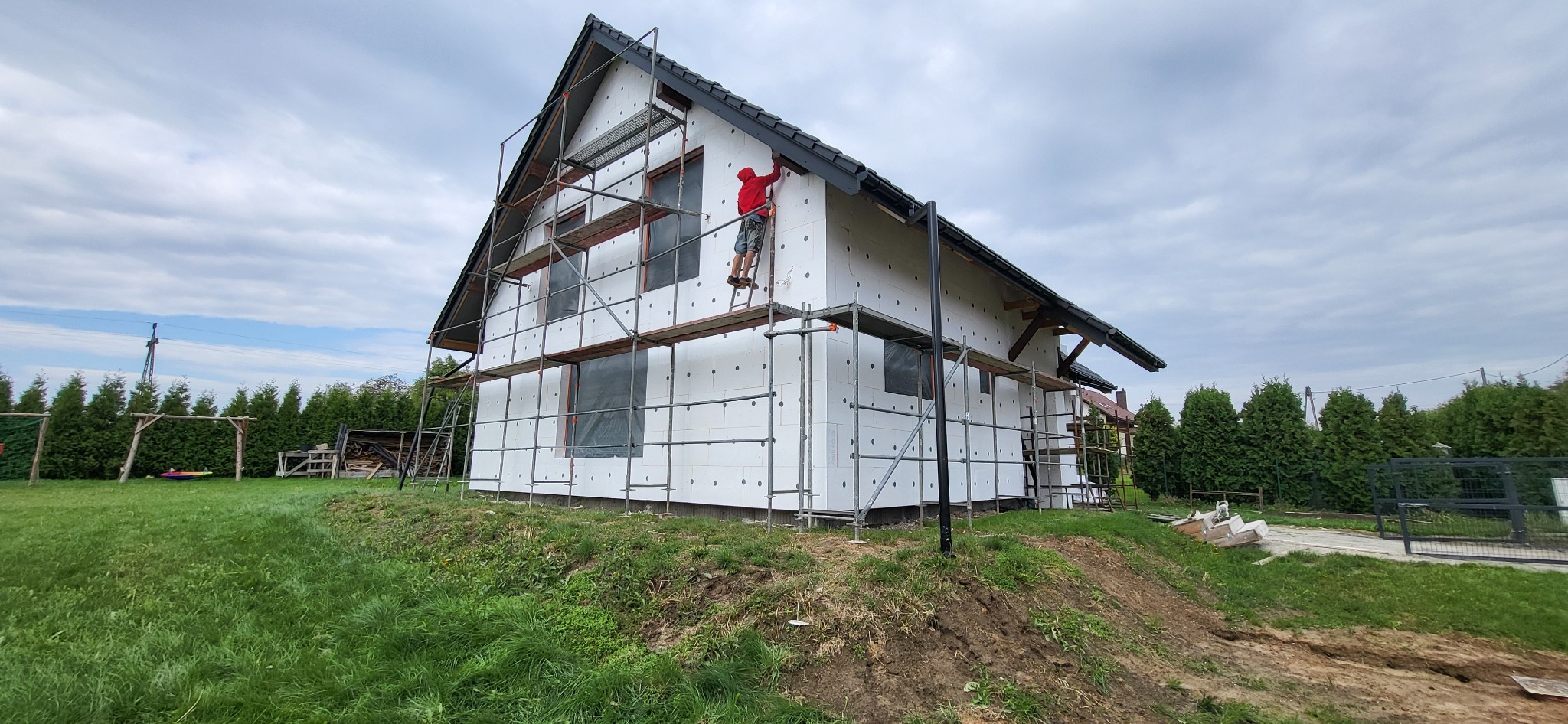 Dom jednorodzinny w trakcie ocieplania: biały styropian na elewacji, rusztowania, pracownik w czerwonej bluzie na drabinie, widok z dołu na tle pochmurnego nieba.