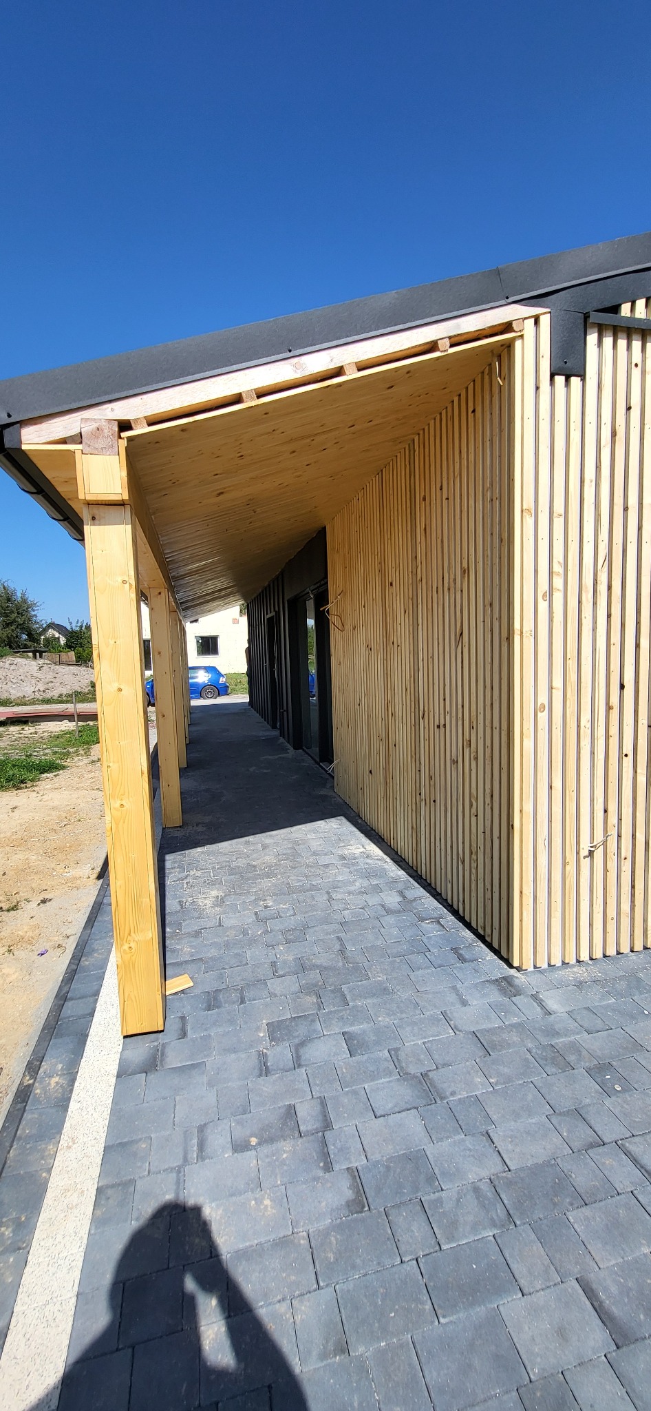 Nowoczesna elewacja budynku z drewnianymi panelami pionowymi i zadaszeniem z surowego drewna, kostka brukowa na podłożu, błękitne niebo w tle.