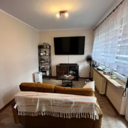 Jasne wnętrze salonu z brązową sofą przykrytą pledem w paski, na podłodze panele imitujące drewno, na ścianie duży telewizor, obok okna z białymi firanami, grzejnik pod oknem.