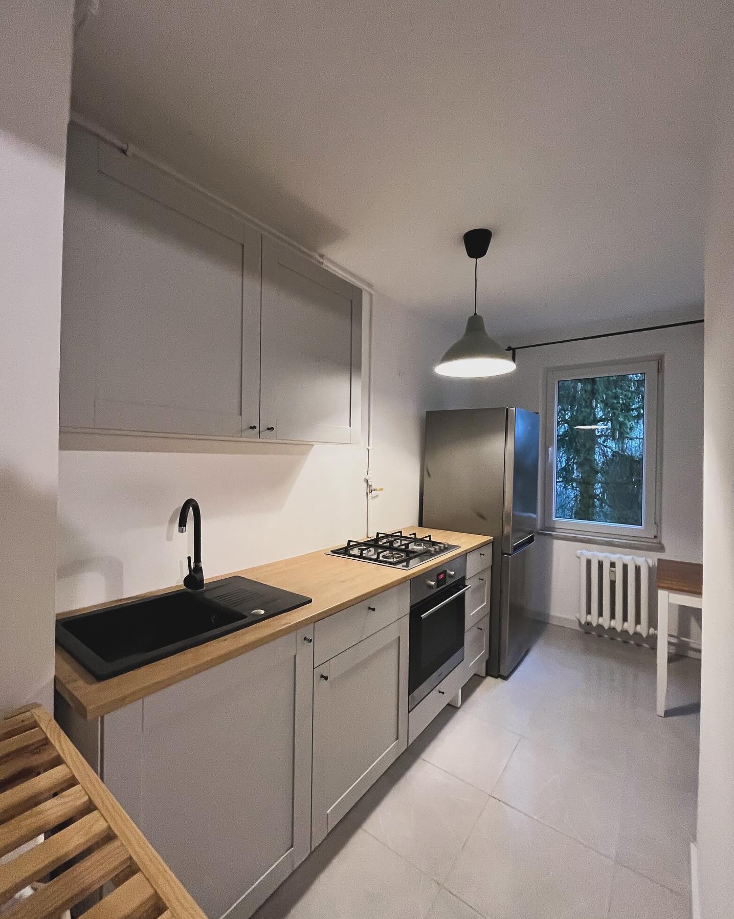 Jasna kuchnia w Miechowie po remoncie: szare szafki, drewniany blat, czarny zlew i bateria, płyta gazowa, piekarnik, lodówka, okno z widokiem na drzewa.