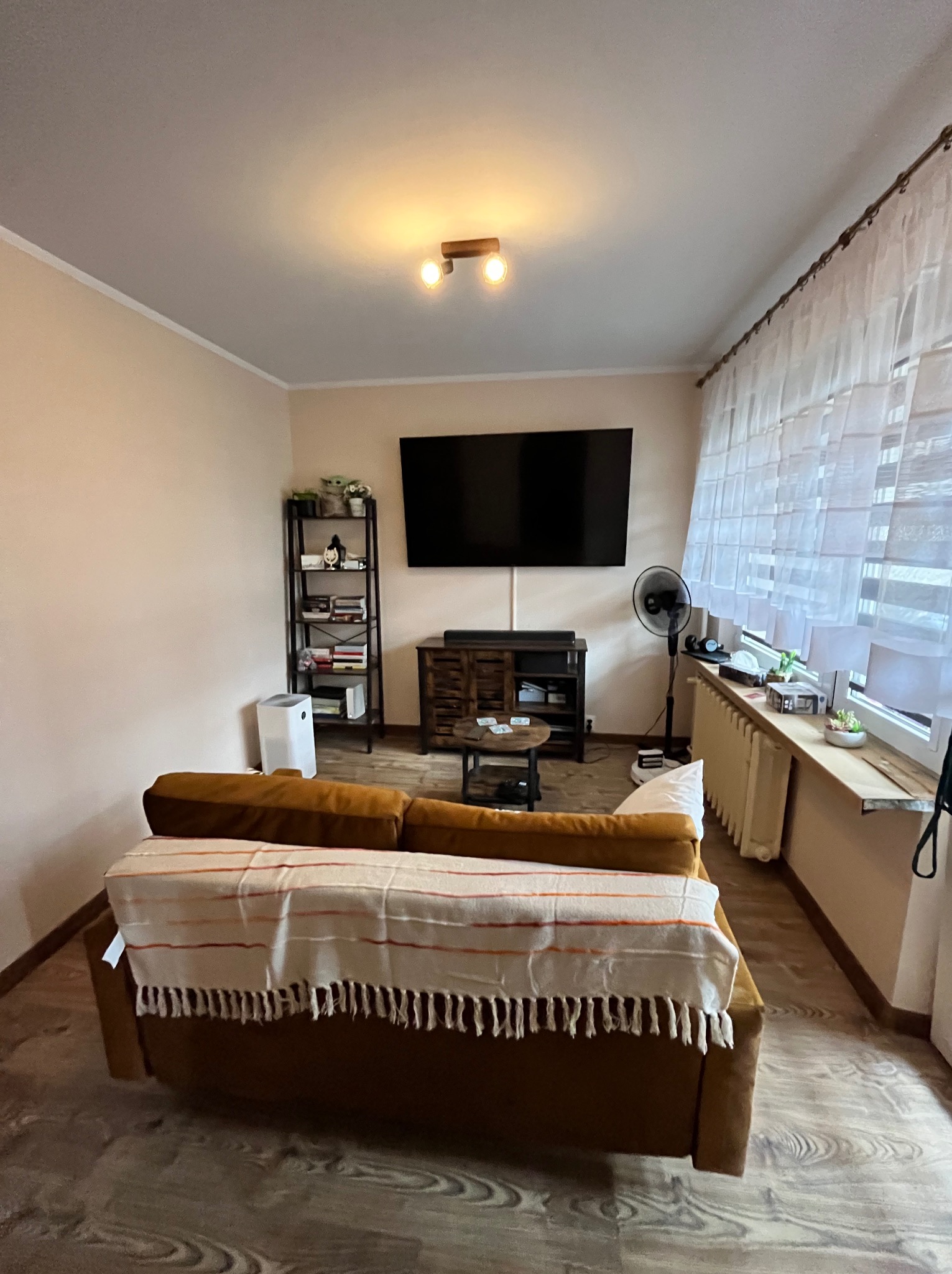 Jasne wnętrze salonu z brązową sofą przykrytą pledem w paski, na podłodze panele imitujące drewno, na ścianie duży telewizor, obok okna z białymi firanami, grzejnik pod oknem.