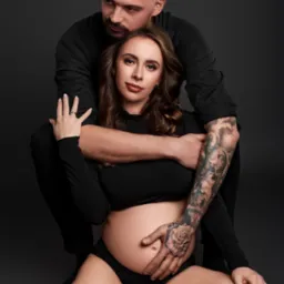 Portret pary: przyszła mama w czarnej bieliźnie i bluzce z długim rękawem, siedząca na podłodze, obejmowana przez partnera z tatuażami na ramieniu, na ciemnoszarym tle.