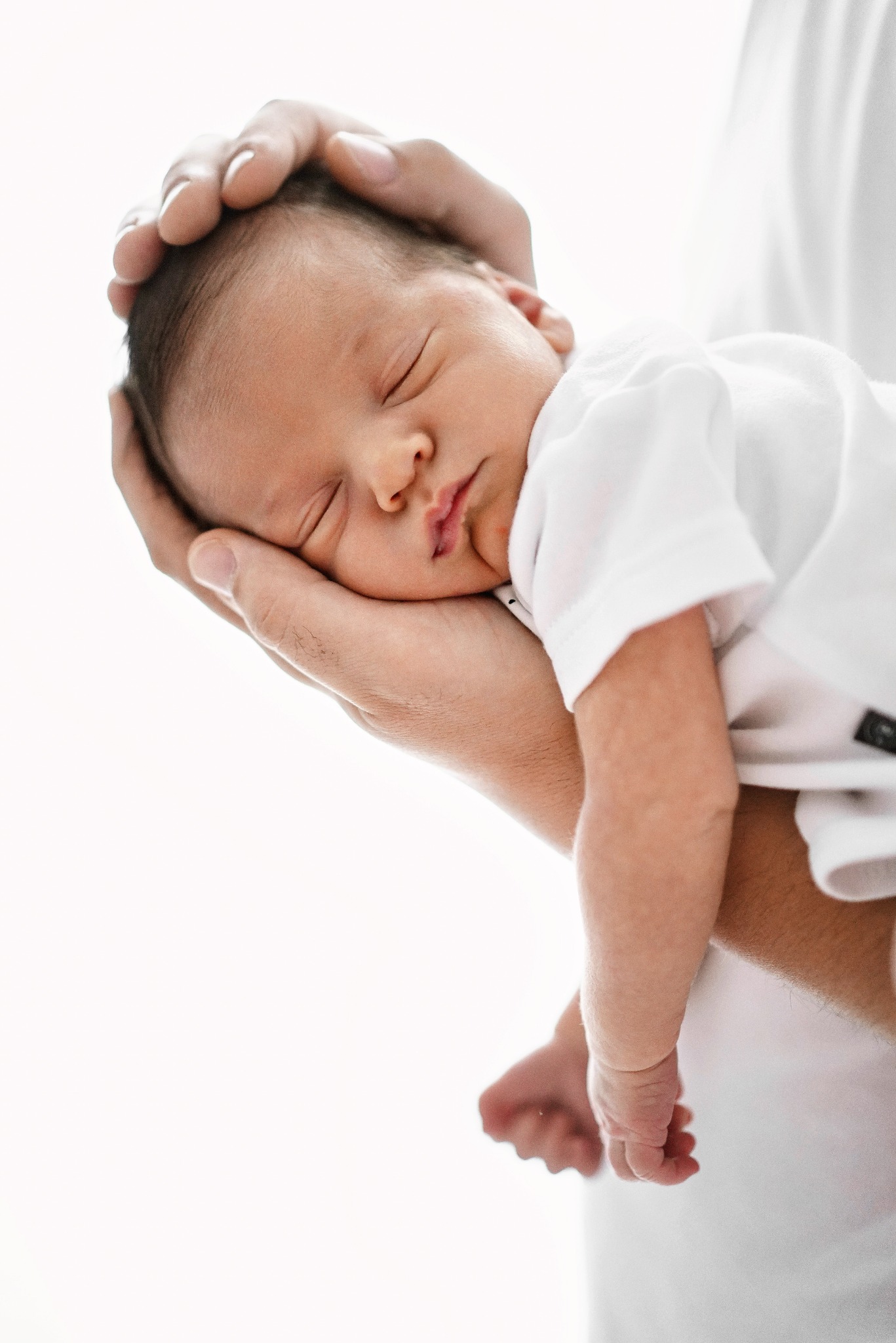 Noworodek w białym ubranku bezpiecznie spoczywa w dłoniach dorosłego, delikatne światło podkreśla jego spokojny sen.