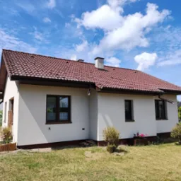 Widok z zewnątrz na ukończony dom jednorodzinny z czerwonym dachem, białymi ścianami i brązowymi oknami w słoneczny dzień z błękitnym niebem i białymi chmurami w tle.