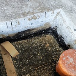 MTM-BUDOWA SPÓŁKA Z OGRANICZONĄ ODPOWIEDZIALNOŚCIĄ - Płyta fundamentowa z izolacją, fragment wykopu z rurą kanalizacyjną i taśmą uszczelniającą. Widoczne ślady obróbki betonu, surowe wykończenie.