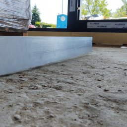 MTM-BUDOWA SPÓŁKA Z OGRANICZONĄ ODPOWIEDZIALNOŚCIĄ - Płyta fundamentowa w trakcie budowy domu modułowego, widoczne okno z czarną ramą i fragment izolacji. Szorstka powierzchnia betonu. Odbiór jakościowy.