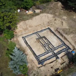 Zbrojenie ław fundamentowych pod budynek dwulokalowy, widok z drona. Czarna folia izolacyjna, wystające pręty zbrojeniowe. W tle drzewa i fragment budynku.