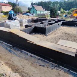 MTM-BUDOWA SPÓŁKA Z OGRANICZONĄ ODPOWIEDZIALNOŚCIĄ - Fundamenty domu jednorodzinnego po izolacji przeciwwilgociowej, zbrojenie wewnątrz, widoczne deski szalunkowe i materiały budowlane w tle.