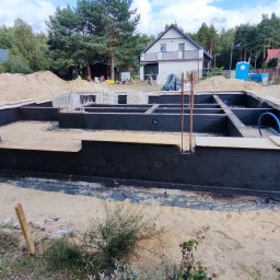 MTM-BUDOWA SPÓŁKA Z OGRANICZONĄ ODPOWIEDZIALNOŚCIĄ - Izolacja przeciwwilgociowa fundamentów domu jednorodzinnego, widoczne czarne ściany fundamentowe, zbrojenie i przygotowany teren pod budowę.